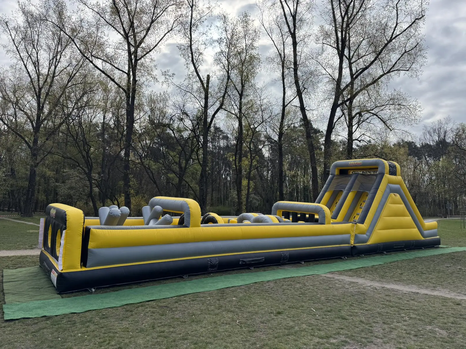 Żółto-czarny dmuchany tor przeszkód 19 m ze zjeżdżalnią na końcu, rozstawiony na trawie w parku